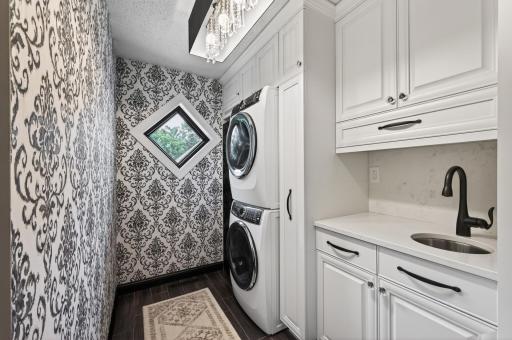 Laundry room featuring custom cabinetry, patterned accent wall, stackable high end washer and dryer, chandelier & most importantly a custom built in steamer closet for all of your dry cleaning delicates -including silk!
