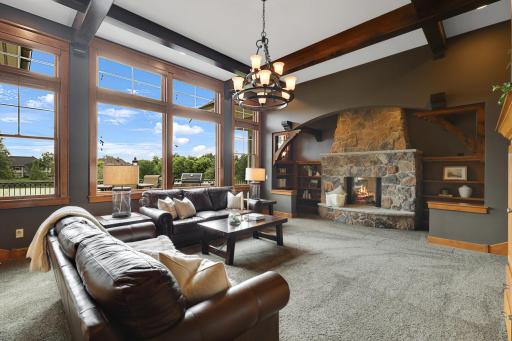 Dramatic great room featuring soaring timber beam-beamed ceilings, a floor to ceiling stone fireplace, custom built-ins, and walls of windows.