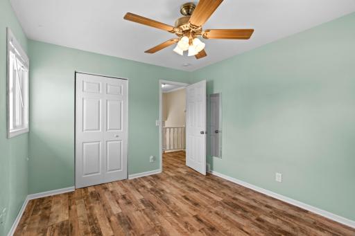 Bright upper-level bedroom at 16860 Foliage Ave W showcasing soft green walls, wood-look plank flooring, an overhead ceiling fan, a paneled closet door, and a clear entryway leading to the hallway staircase.