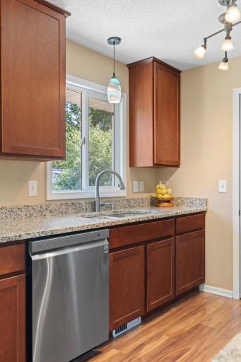 Kitchen detail at 16860 Foliage Ave W highlighting speckled stone-look countertops, an undermount stainless steel sink with pull-down faucet, an adjacent dishwasher, warm wood cabinetry, and a bright window overlooking the yard.
