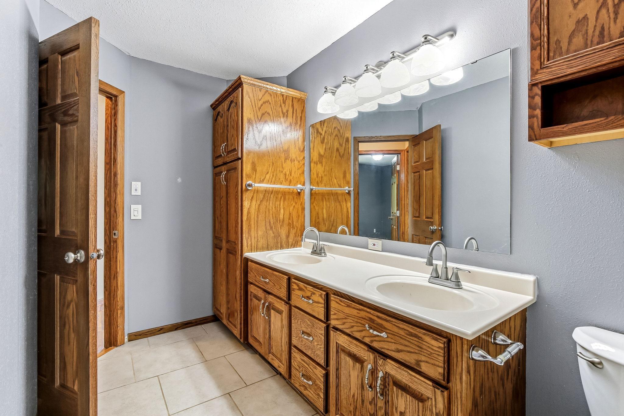 Well-appointed bathroom with a double vanity, extensive cabinet storage, and tile flooring. The roomy layout offers excellent functionality, making mornings easier while providing plenty of space for organization.