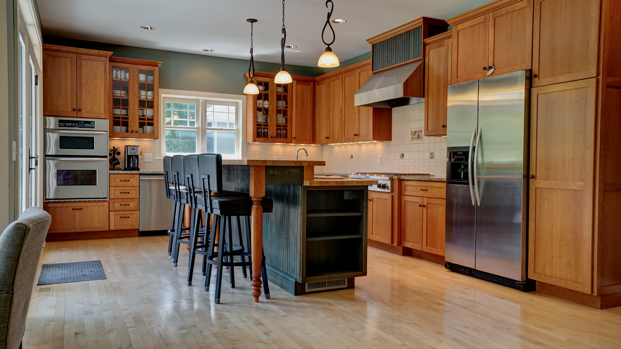 An expansive 18x15 kitchen featuring custom cherry cabinetry. An 8-foot center island anchors the kitchen, complete with a dual-height butcher block counter that seats four. KitchenAid double wall-ovens and a heavy-duty side-by-sde refrigerator.