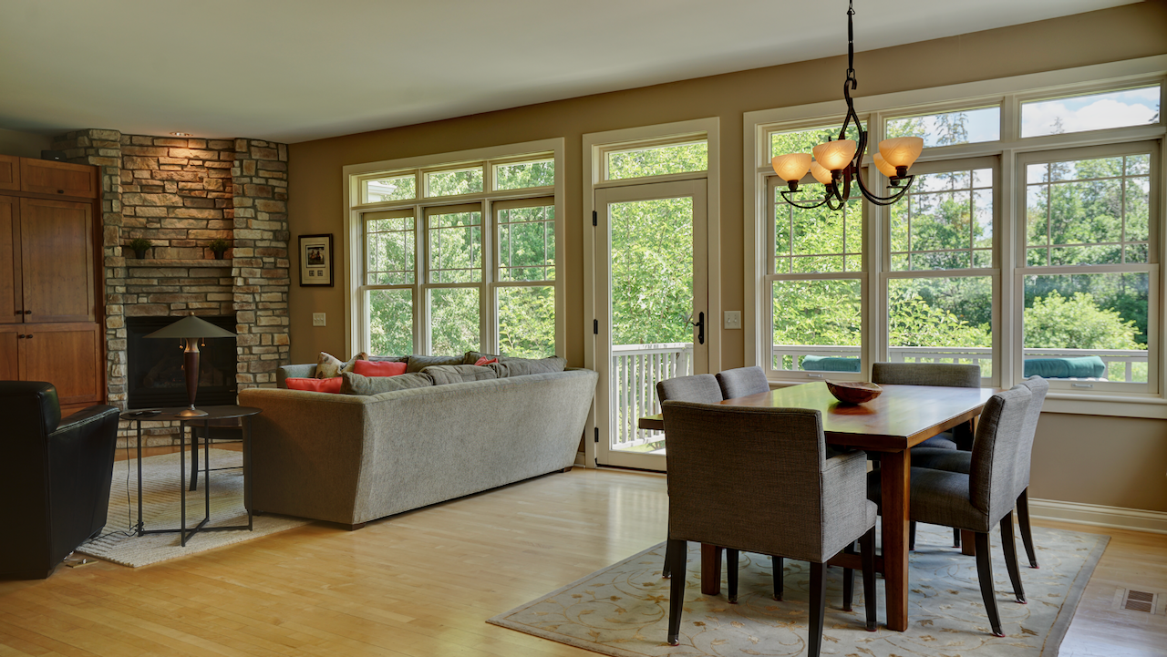 The layout flows smoothly from the dining space into the living area, lined by a wall of windows. A second glass door opens directly to the wrap-around deck, framing private, preserve-like views over the nearly one-acre lot.