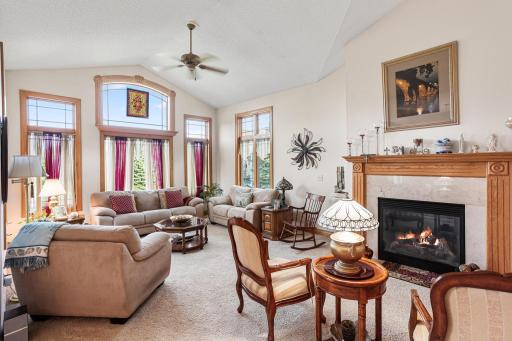 The spacious living room features vaulted ceilings, a gas fireplace with wood mantel, ceiling fan, and expansive windows that bring in abundant natural light and showcase backyard views.