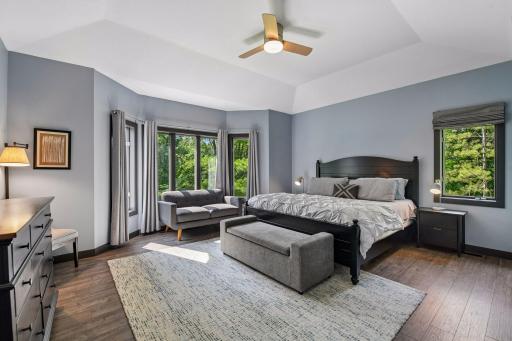 A spacious primary bedroom with a tray ceiling, ceiling fan, and a wall of windows overlooking the trees.