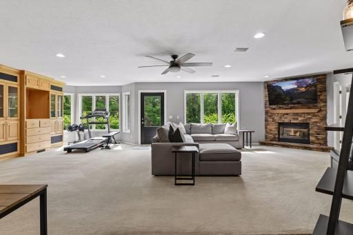 The finished walkout lower level offers a spacious family room with a stone fireplace, recessed lighting, a ceiling fan, and large windows bringing in natural light and wooded views.