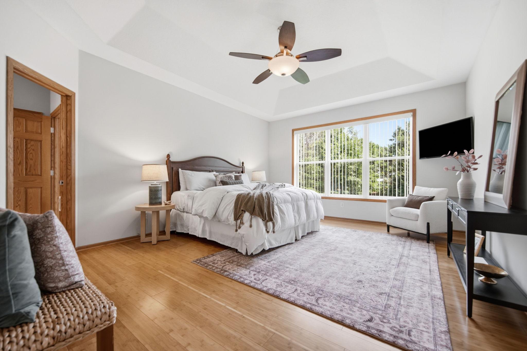 The spacious primary bedroom is conveniently located on the main floor & features a tray ceiling, abundant natural light, & natural wood flooring. The room easily accommodates a king-size bed as seen here, highlighting its generous proportions.