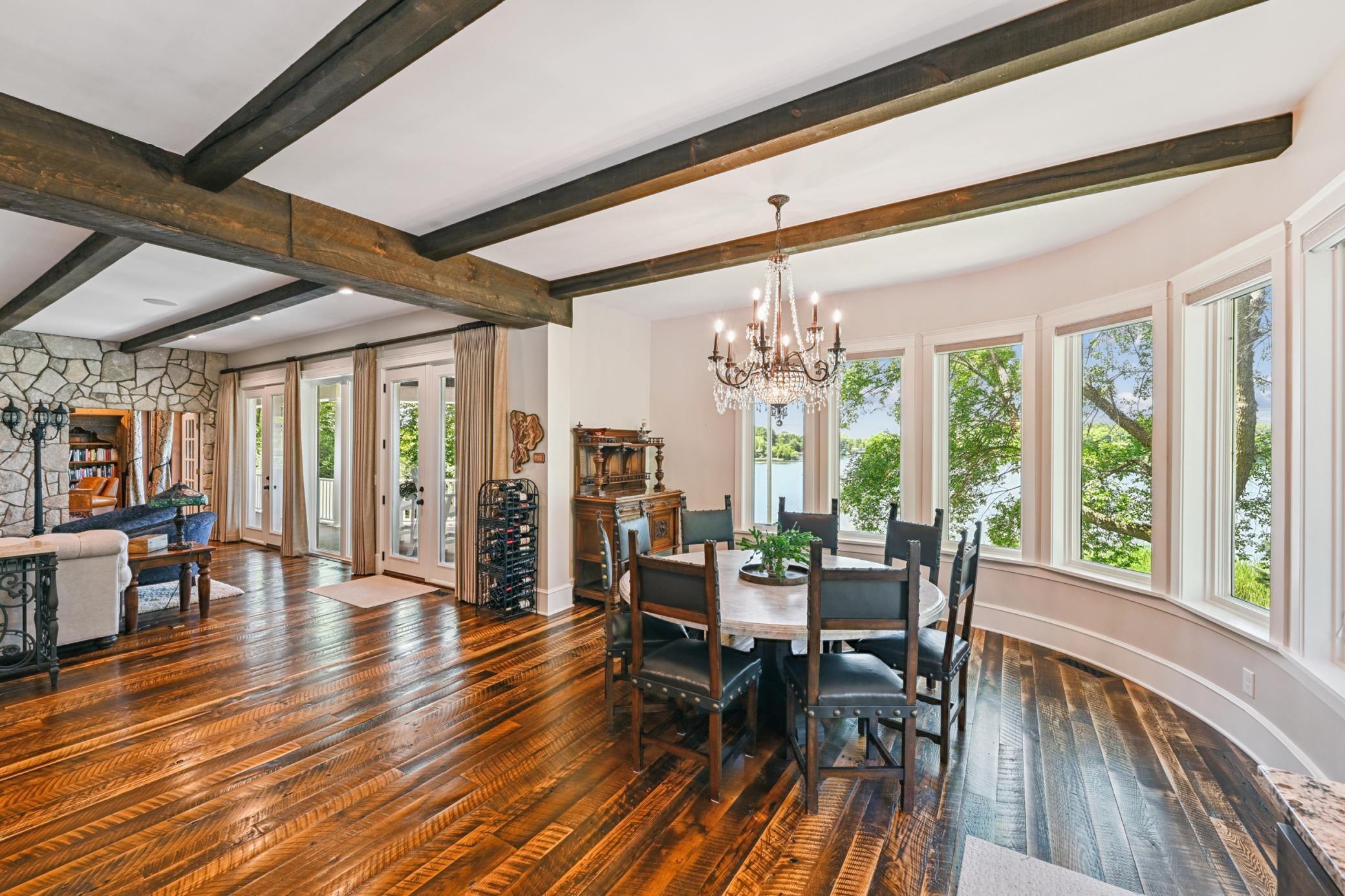 Elegant dining area framed by a curved wall of lakeside windows with panoramic water views, rich hardwood floors, exposed ceiling beams, and a sparkling chandelier, perfect for memorable meals by the lake.