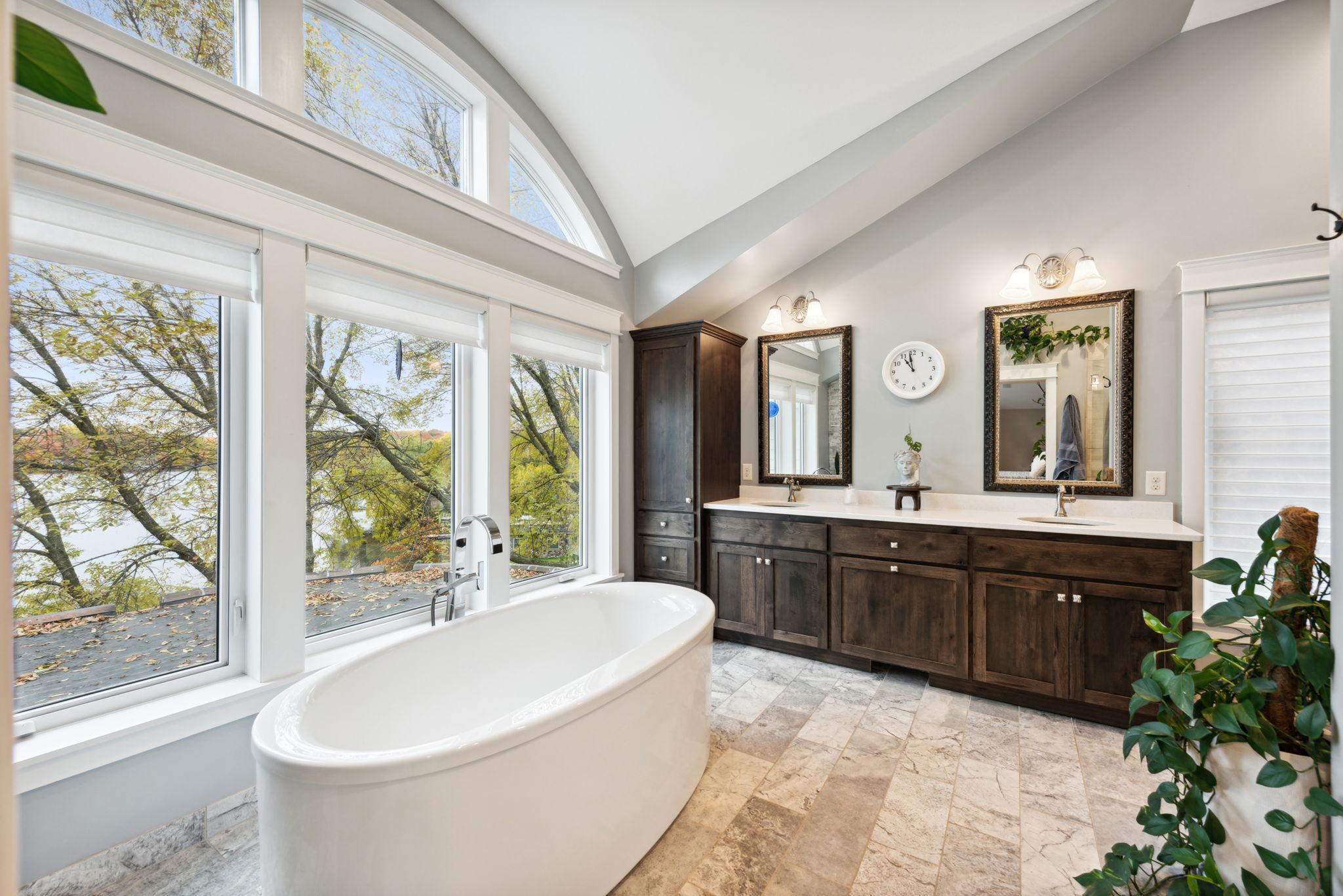 Relax in the spa-like primary bath featuring vaulted ceilings, a walk-in shower, a soaking tub with scenic lake views, dual vanities, and custom cabinetry.