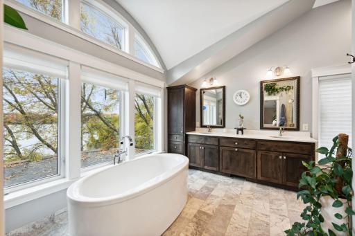 Relax in the spa-like primary bath featuring vaulted ceilings, a walk-in shower, a soaking tub with scenic lake views, dual vanities, and custom cabinetry.