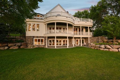 This lakeside view highlights the homeâs elegant architecture with three levels of covered outdoor living, natural stonework, and beautifully landscaped grounds just steps from the waterâs edge.