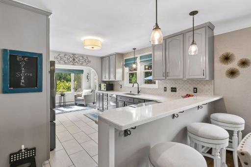 Quartz breakfast bar connects the dining room to the kitchen, with space for bar seating while you prepare meals. Two-toned painted gray cabinetry with white countertops, black stainless appliances, and sitting nook in rear.