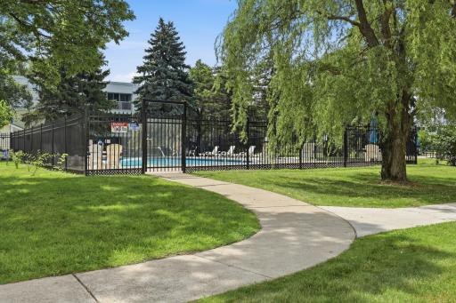 A winding sidewalk leads through lush green lawns toward a sparkling community pool, creating a sense of calm and quiet retreat.