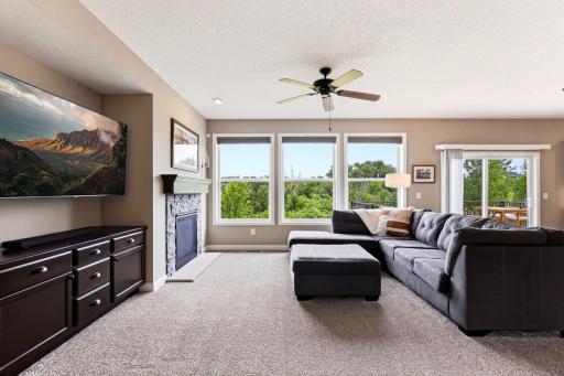 The great room centers on a stacked stone gas fireplace, with a wall of windows overlooking the green space beyond the deck.