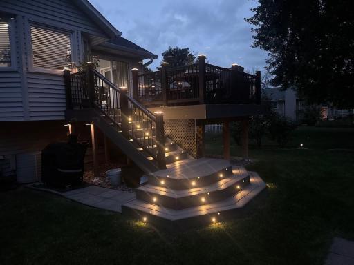 Evening photo of the 16 x 12 maintenance free deck glowing with color changing LED stair lighting. The lights, controlled by remote, shift hues and pulse to music, creating a vibrant, inviting outdoor space leading to the fire pit and gazebo hot tub.