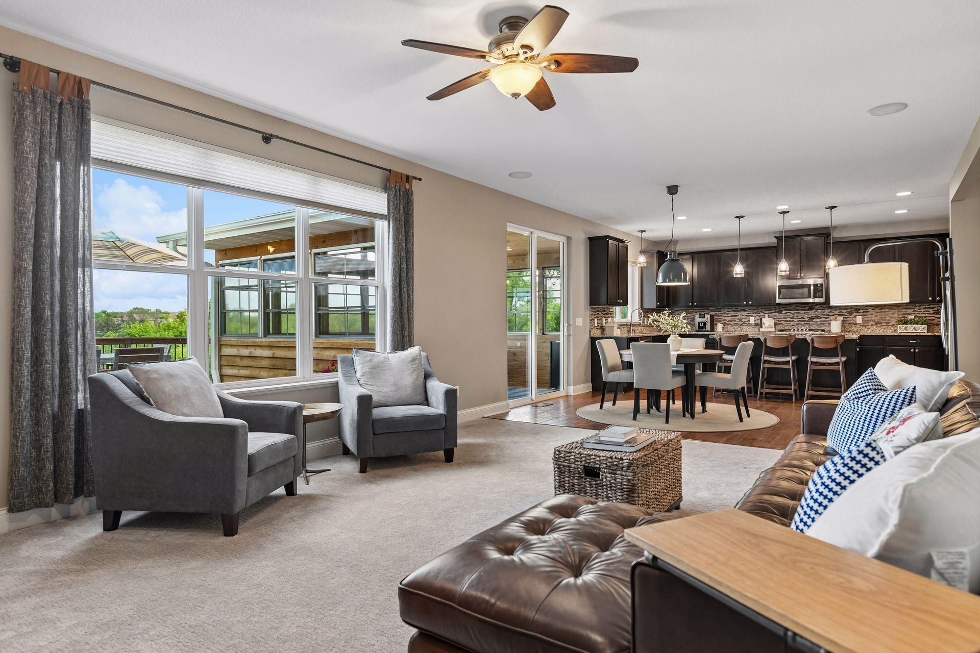 Open-concept main level with the living area flowing into the dining space and kitchen, lit by a ceiling fan and a wall of windows.