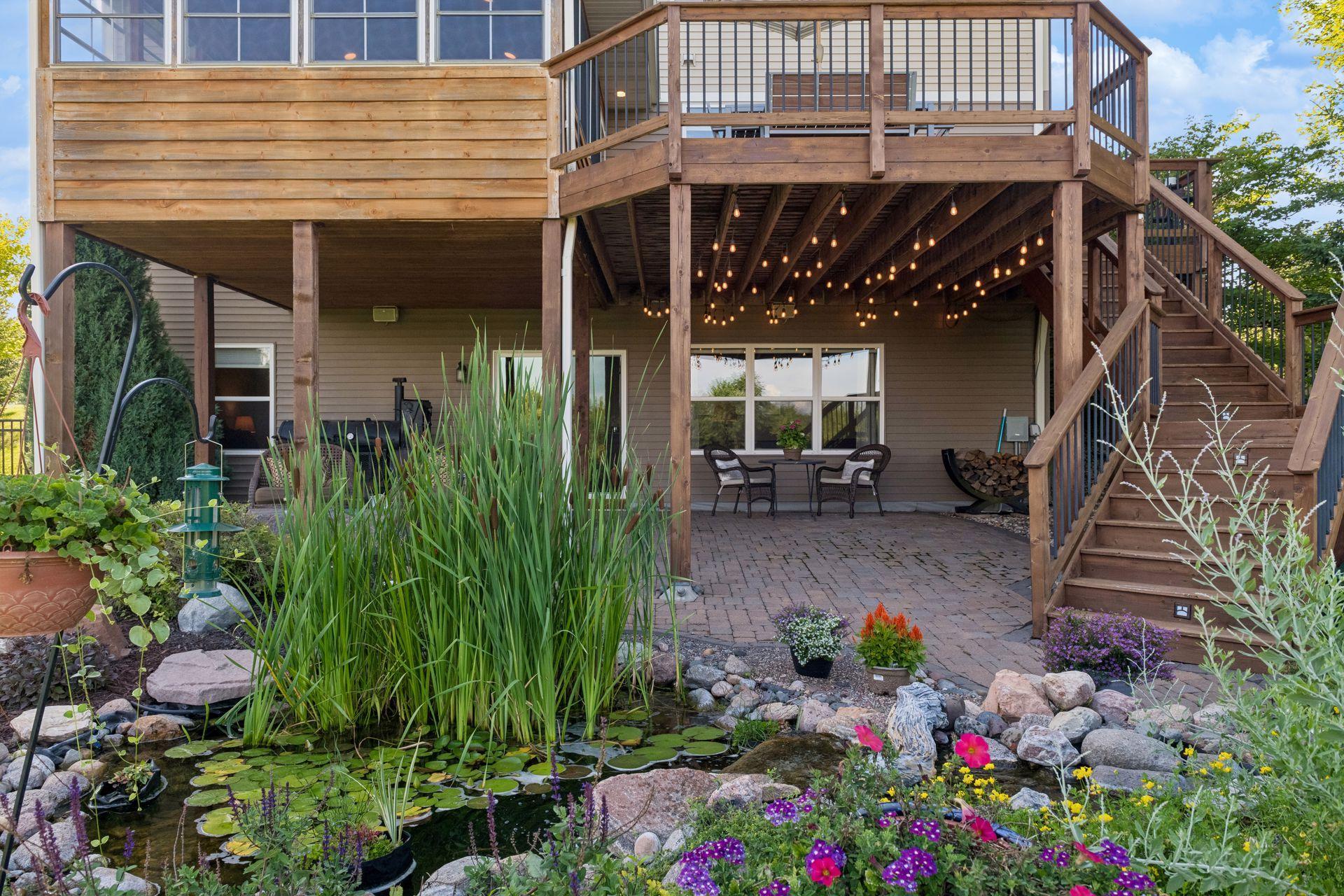 Covered patio beneath the deck with string lighting, overlooking the water feature and landscaped beds.