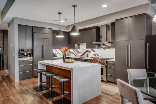 The chefâs kitchen features a striking waterfall island with seating, Cambria countertops and statement backsplash, Wolf and Sub-Zero appliances, Miele built-in coffee maker, and gorgeous custom cabinetry.