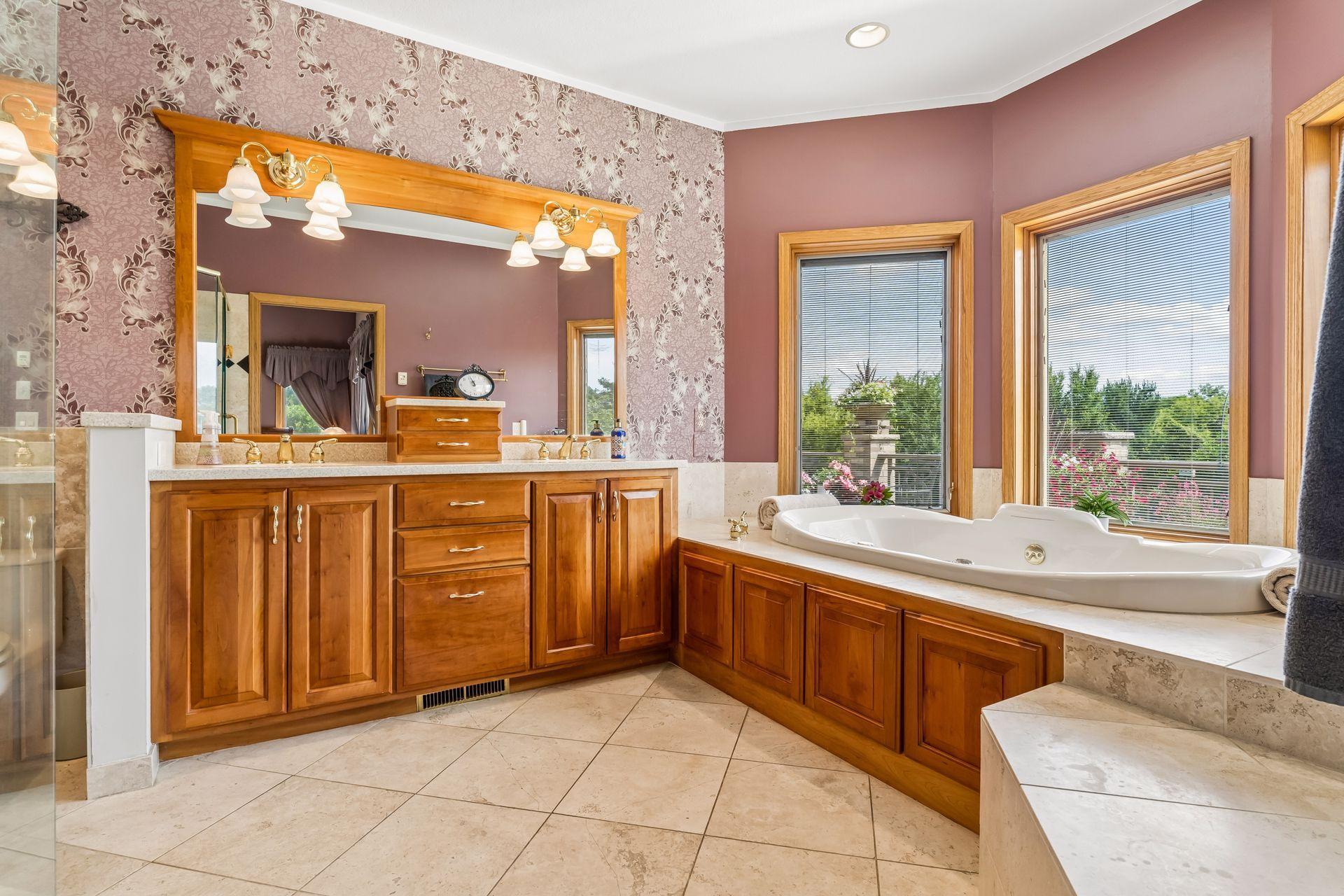 The expansive primary bathroom offers a spa-like retreat with a Jacuzzi tub, elegant cherry wood double vanity, custom heated tile floors, abundant natural light from the windows, and recessed lighting throughout.