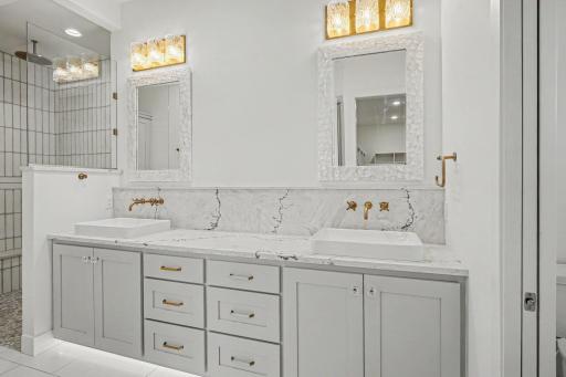 Spa-inspired owner's bath featuring dual quartz vanities, custom cabinetry, designer lighting, and elegant gold fixtures.