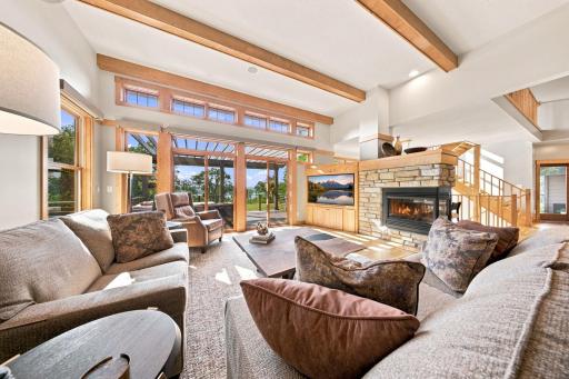 The stunning great room is anchored by a striking three-sided fireplace, motorized window shades, and expansive glass doors leading to maintenance-free decking overlooking the river.