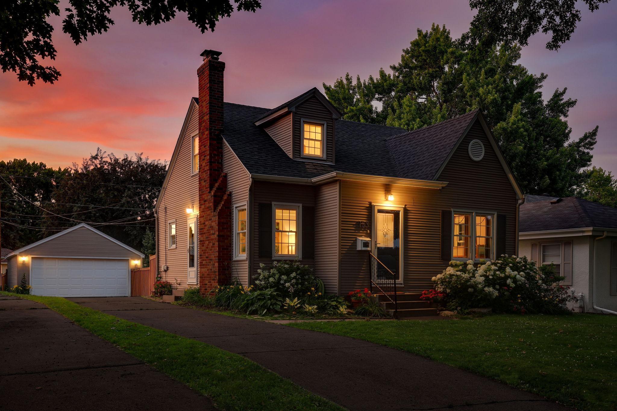 Twilight with of property from the southwest, showing all of the warm welcome inside the house and two car garage at end of driveway and surrounding landscaping.