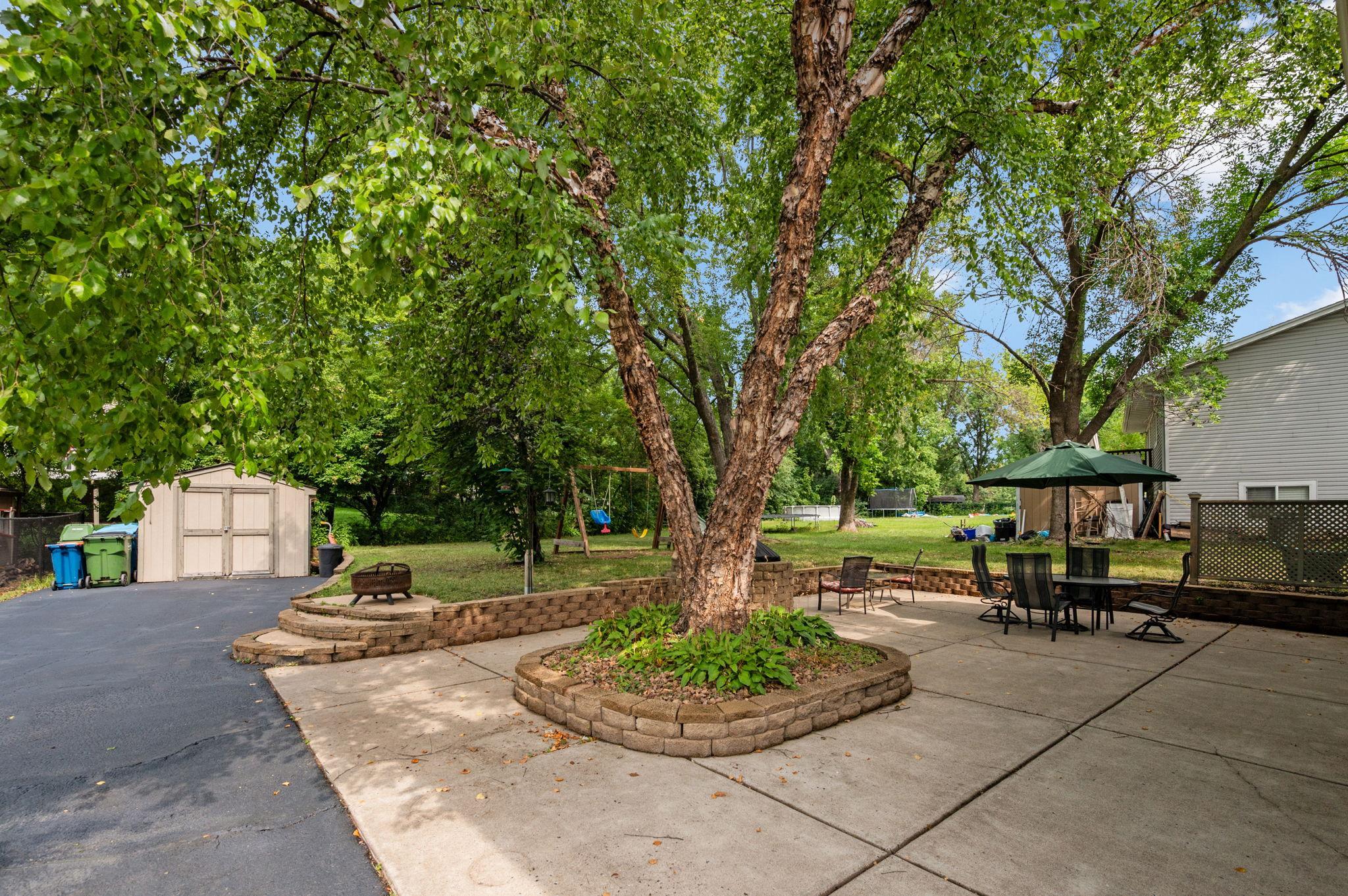 Expansive paved patio surrounded by mature shade trees, complete with a center garden island and outdoor seating space