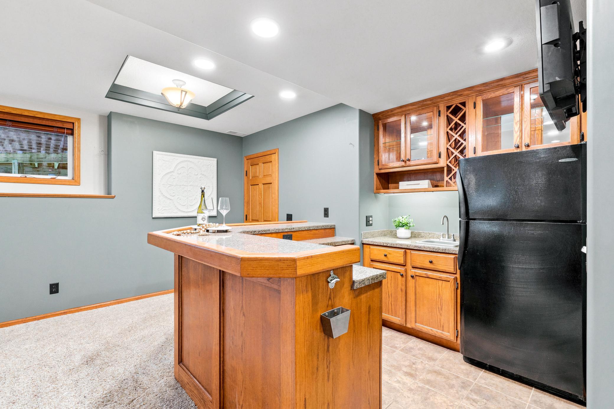 Entertain with ease at the custom wet bar featuring rich oak cabinetry, granite-look countertops, glass display cabinets, built-in wine rack, refrigerator, sink, and expansive serving peninsula.