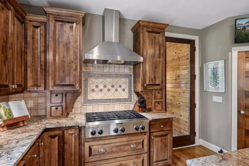 A stunning cooking station featuring a gas cooktop, large stainless hood vented to the exterior, and a decorative tile mosaic that adds beautiful visual interest.