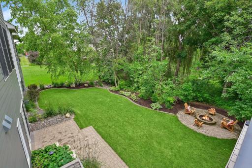 A beautiful view from the deck reveals a private backyard retreat with a paver firepit, mature trees, shrubs, and perennials, all accented by concrete curbed edging and automatic landscape lighting.