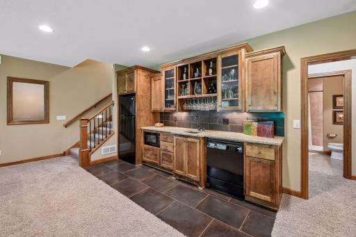 The lower-level family room features a wet bar with glass-front maple cabinetry, under-cabinet lighting, granite counters, a dishwasher, fridge, and microwave, along with practical tile flooring.