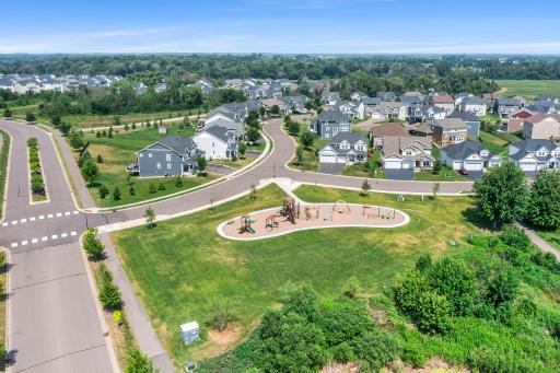 High aerial view framing the neighborhood play area, surrounding green lawn, paved sidewalks, and tree-lined streets.