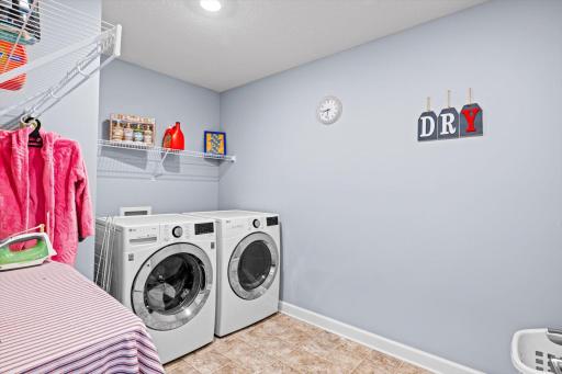 Convenient upper-level laundry room with front-loading washer and dryer, vinyl tile flooring, and built-in overhead wire shelving.
