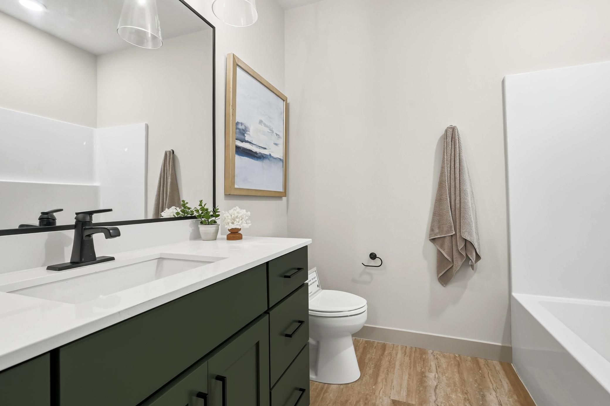 This minimilast bathroom features a sleek black faucet over a white countertop, complemented by a beige towel and a small potted plant. Soft, ambient lighting enhances the clean, modern aesthetic, with neutral walls and a simple, ele