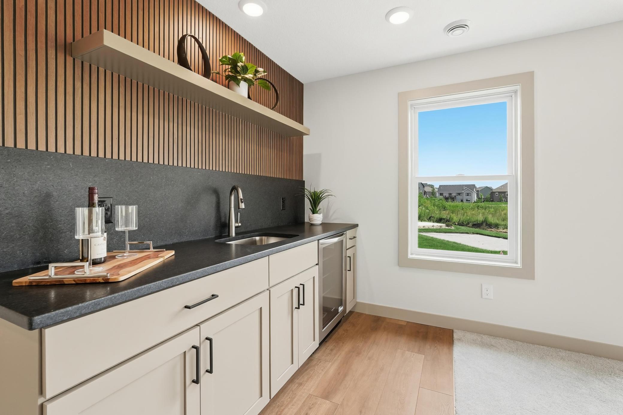 Stylish lower-level wet bar equipped with slatted-wood wall paneling, a floating shelf, dark quartz countertops, built-in beverage fridge, and bright natural views.