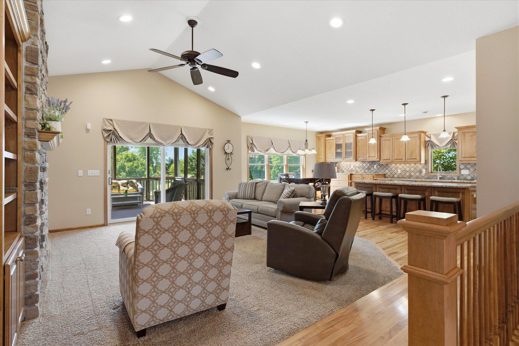 Open-concept great room seamlessly connects the living area, dining space, and kitchen, showcasing vaulted ceilings, recessed lighting, a floor-to-ceiling stone fireplace, and sliding glass doors leading to the screened porch.