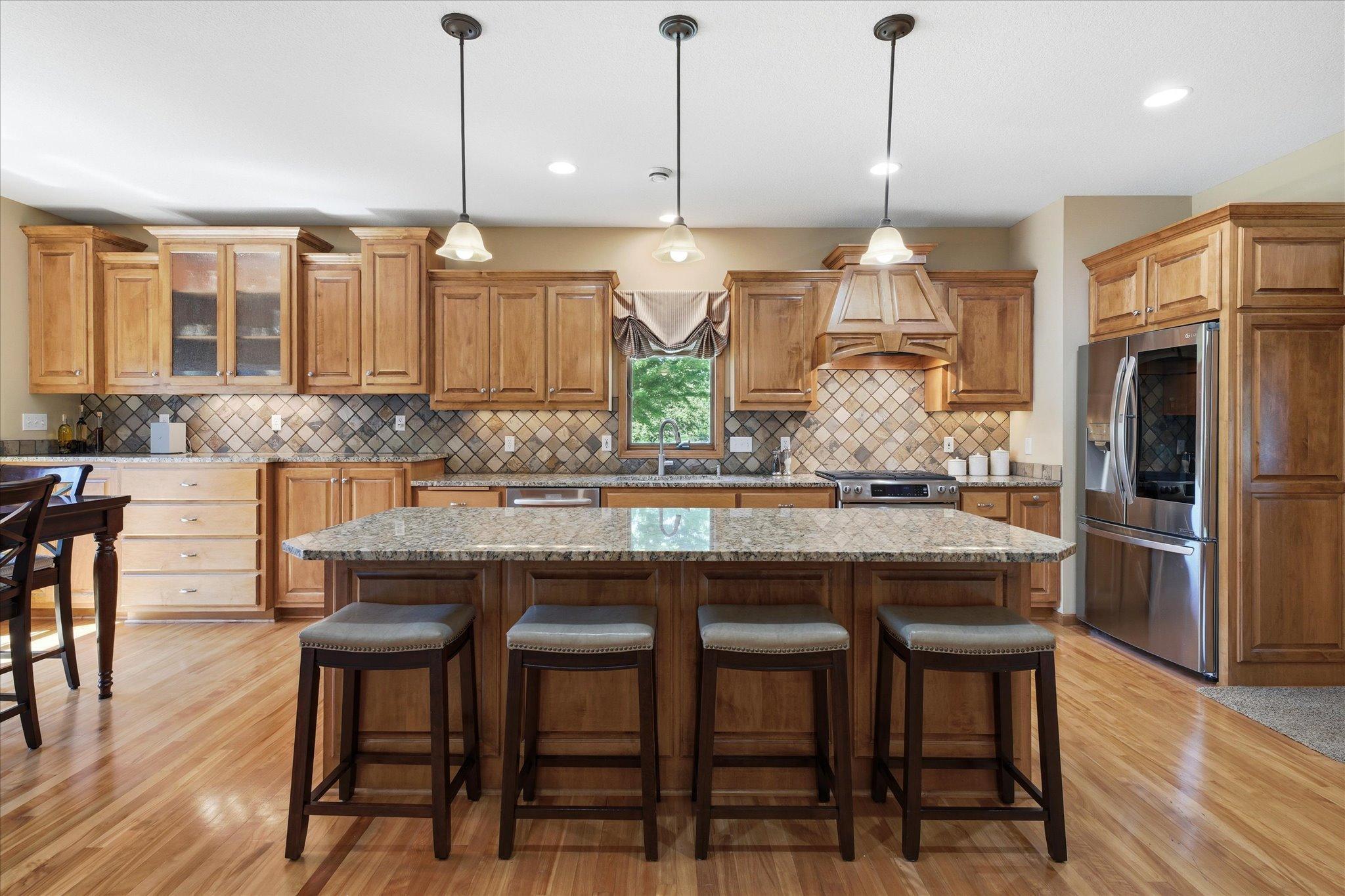 The heart of the home features an oversized granite island, custom maple cabinetry, updated stainless steel refrigerator, gas range, tile backsplash, and abundant prep space for everyday living and entertaining.