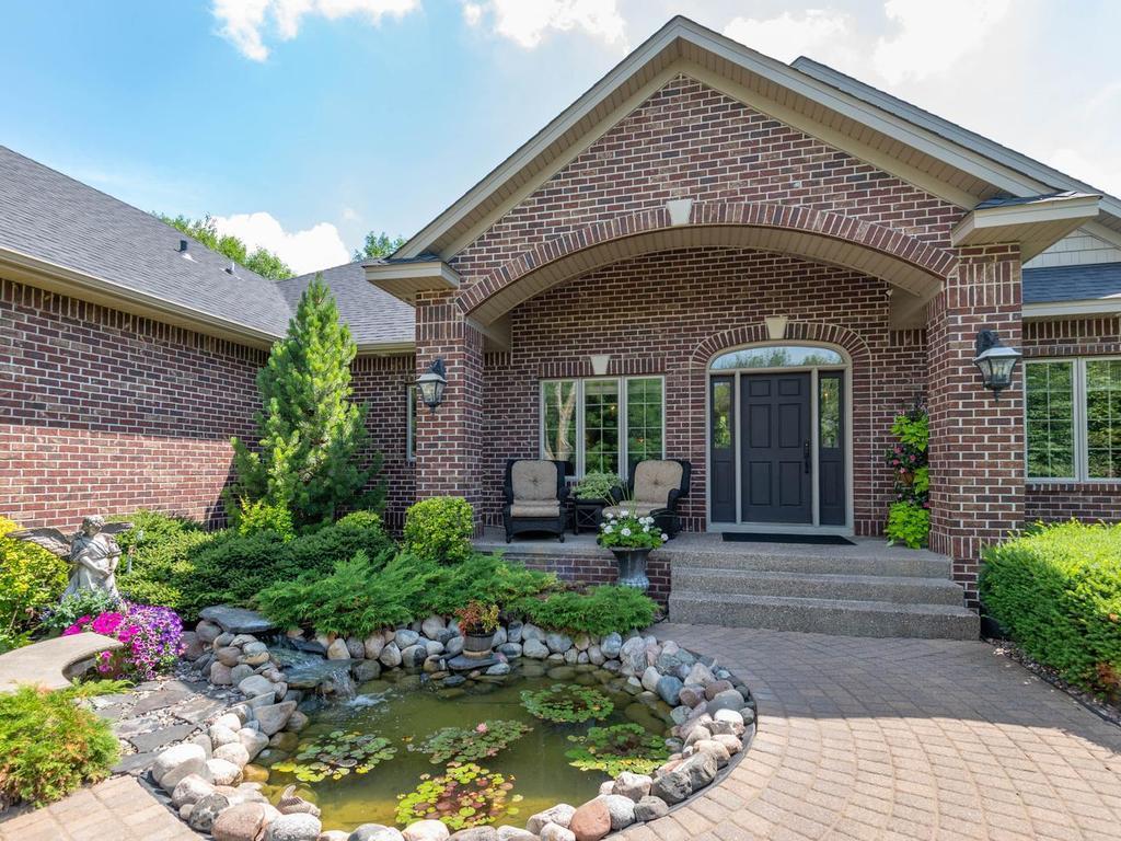 Welcoming covered front entry framed by brick architecture, mature landscaping, curved paver walkways, and a decorative pond with a waterfall.