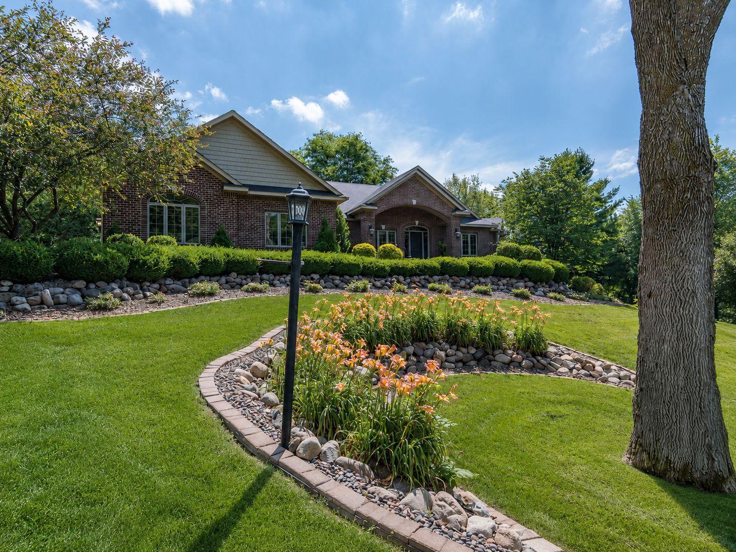 Beautifully landscaped front yard with a curved stone-edged garden beds, flowering perennials, mature trees, and a manicured lawn leading to the front entry.