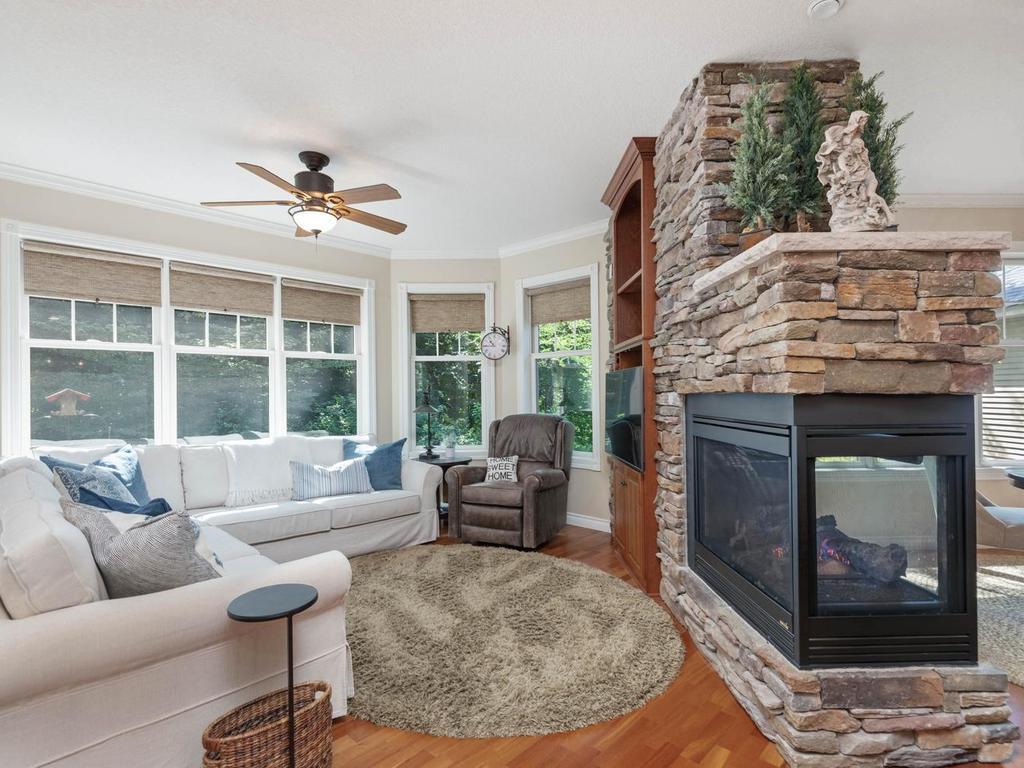 Window-filled hearth room features hardwood flooring, ceiling fan, built-in cabinetry, and a striking 3-sided stone gas fireplace shared with the kitchen and informal dining area.