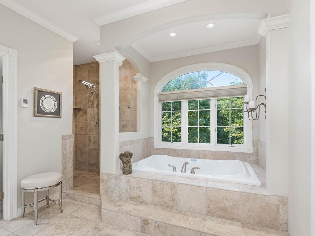 Primary bath includes a tiled jetted whirlpool tub beneath an arched window, separate walk-in tiled double shower, architectural columns, recessed lighting, and wooded views.