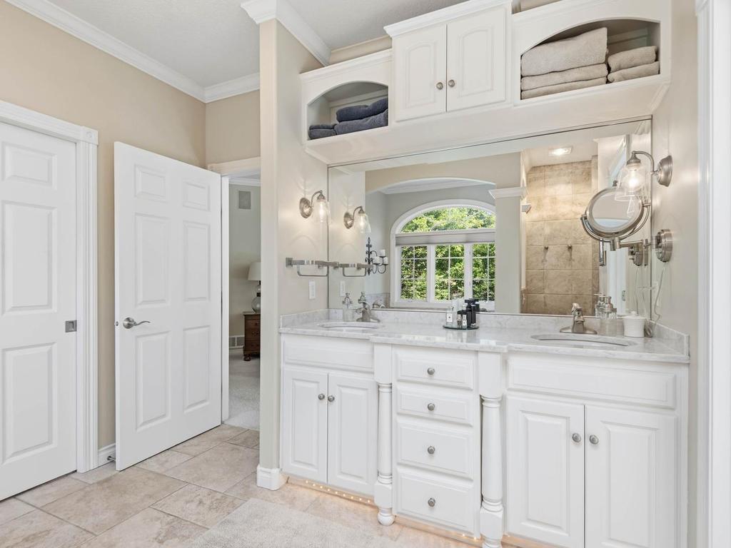 Primary bath features a dual-sink vanity, tile flooring, built-in upper storage, decorative lighting, and an arched opening leading to the soaking tub area with wooded views.