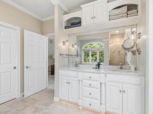 Primary bath features a dual-sink vanity, tile flooring, built-in upper storage, decorative lighting, and an arched opening leading to the soaking tub area with wooded views.