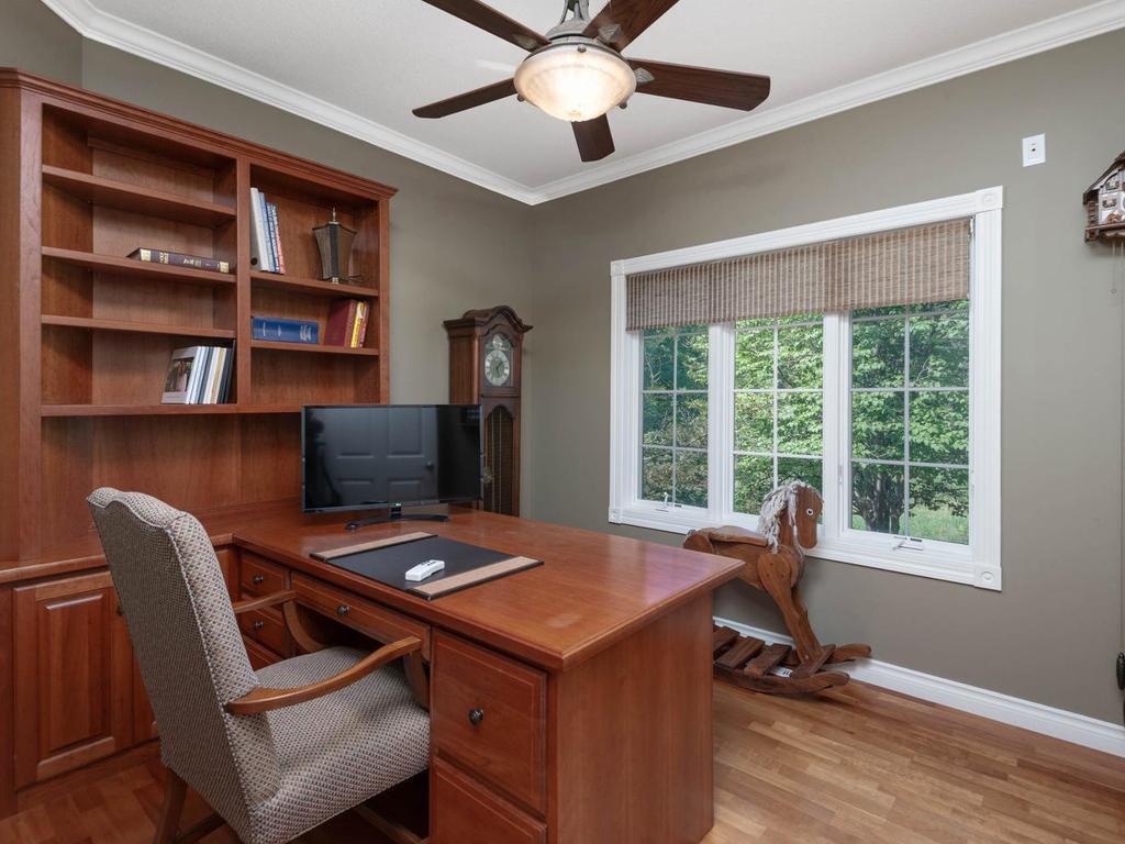 Main level office features hardwood flooring, crown molding, ceiling fan, expansive built-in desk and shelving, and a large window overlooking the wooded setting.