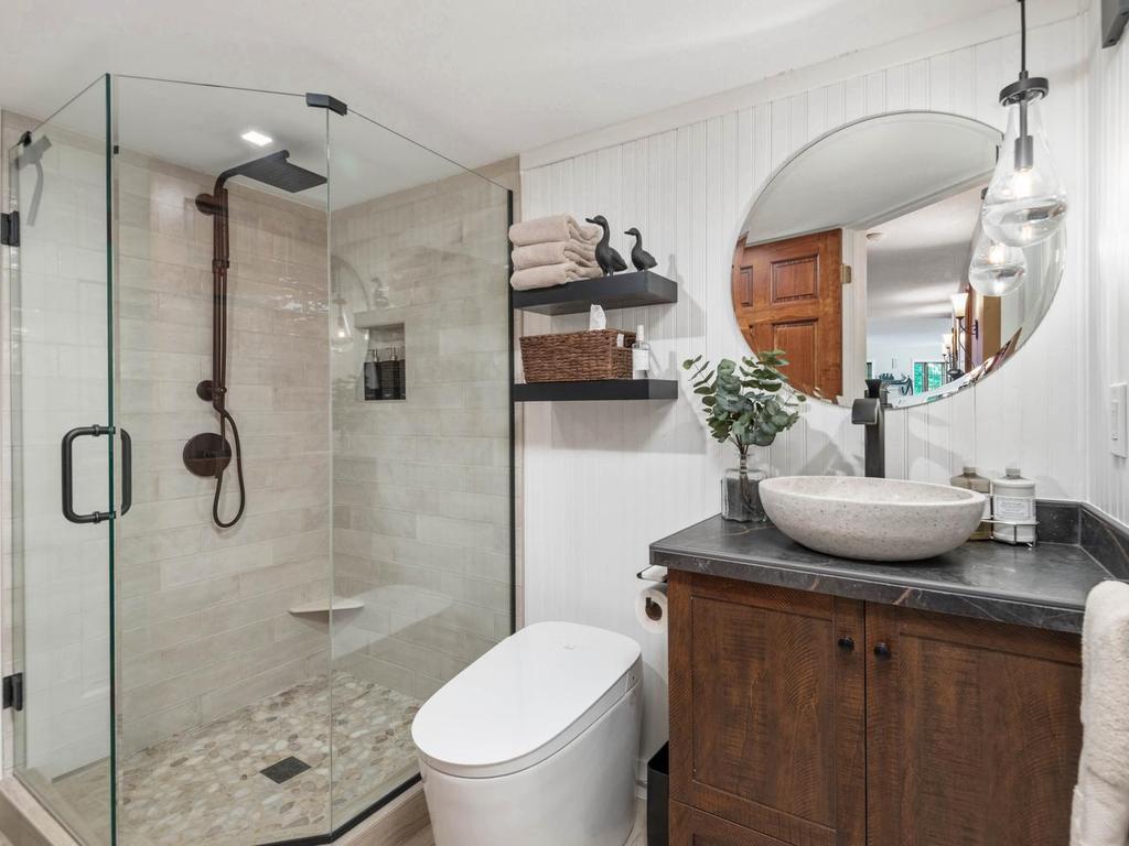 Beautiful lower level ¾ bath features a frameless glass walk-in shower with a rainfall showerhead, tiled surround and floor, vessel sink vanity, floating shelves, and contemporary lighting.