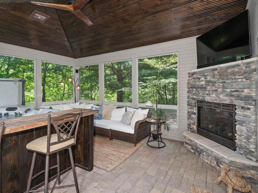 Screened porch includes a striking stone gas FP, creating an inviting focal point alongside the hot tub, paver floor, vaulted wood ceiling, and wooded views.