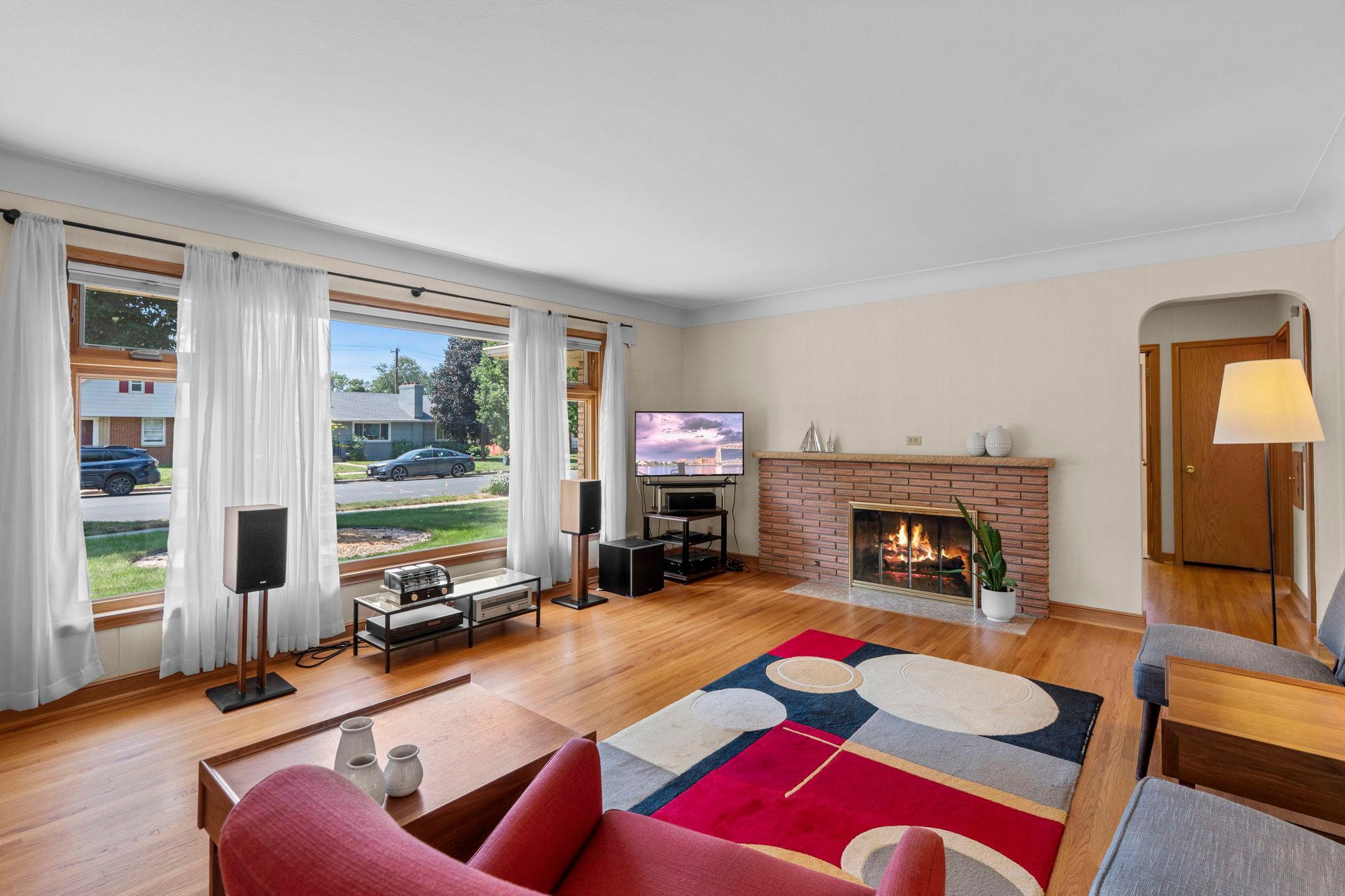The welcoming living room is anchored by a classic brick wood-burning fireplace, beautiful hardwood floors, and oversized windows that fill the space with natural light.