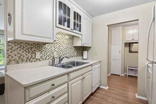 Crisp clean kitchen with vinyl flooring, tile back splash, white cabinetry with glass display cabinet, & white appliances.
