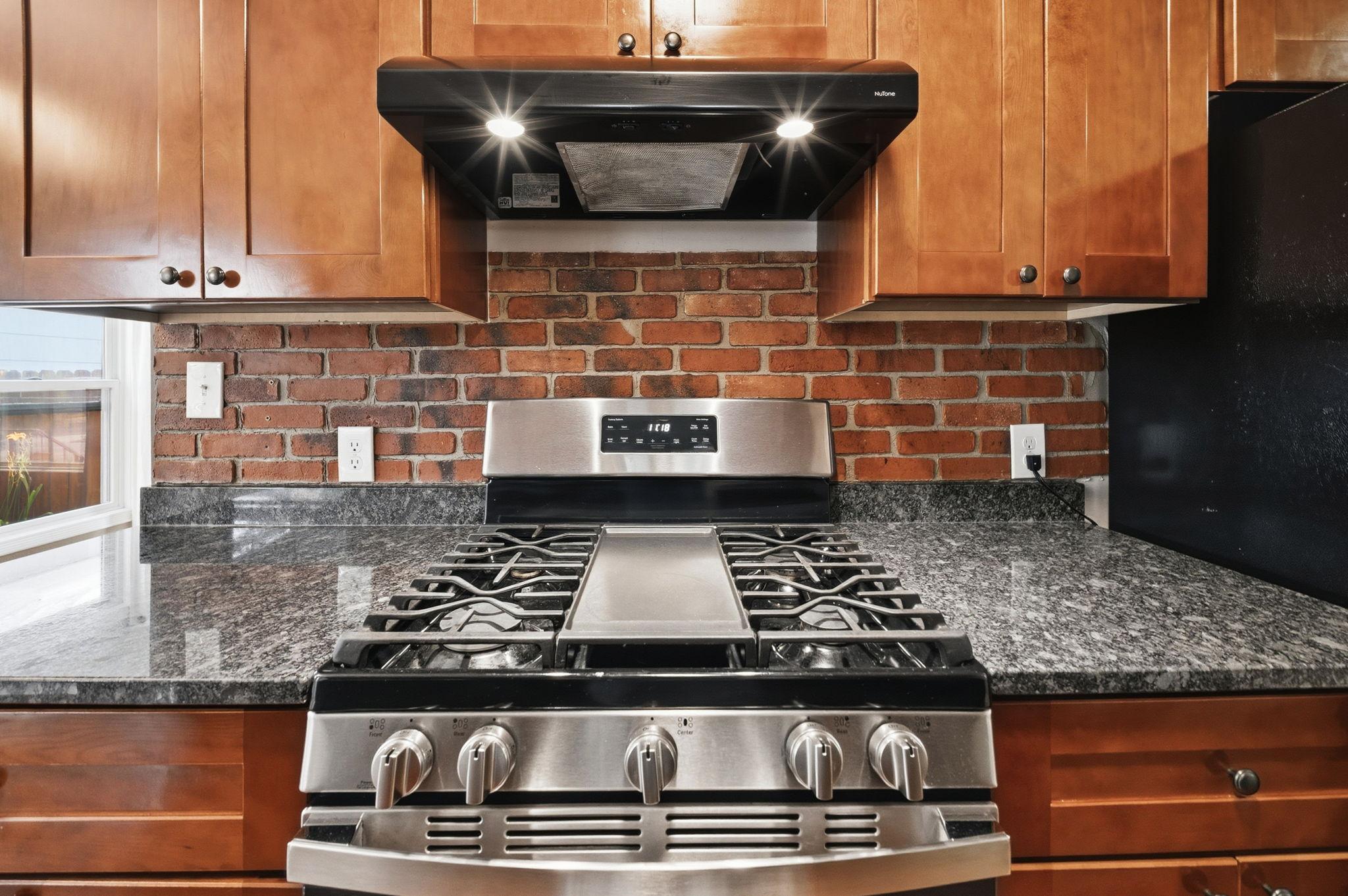 A five burner gas range set against a brick backdrop. This is where Sunday sauce simmers for hours and nobody minds the wait.