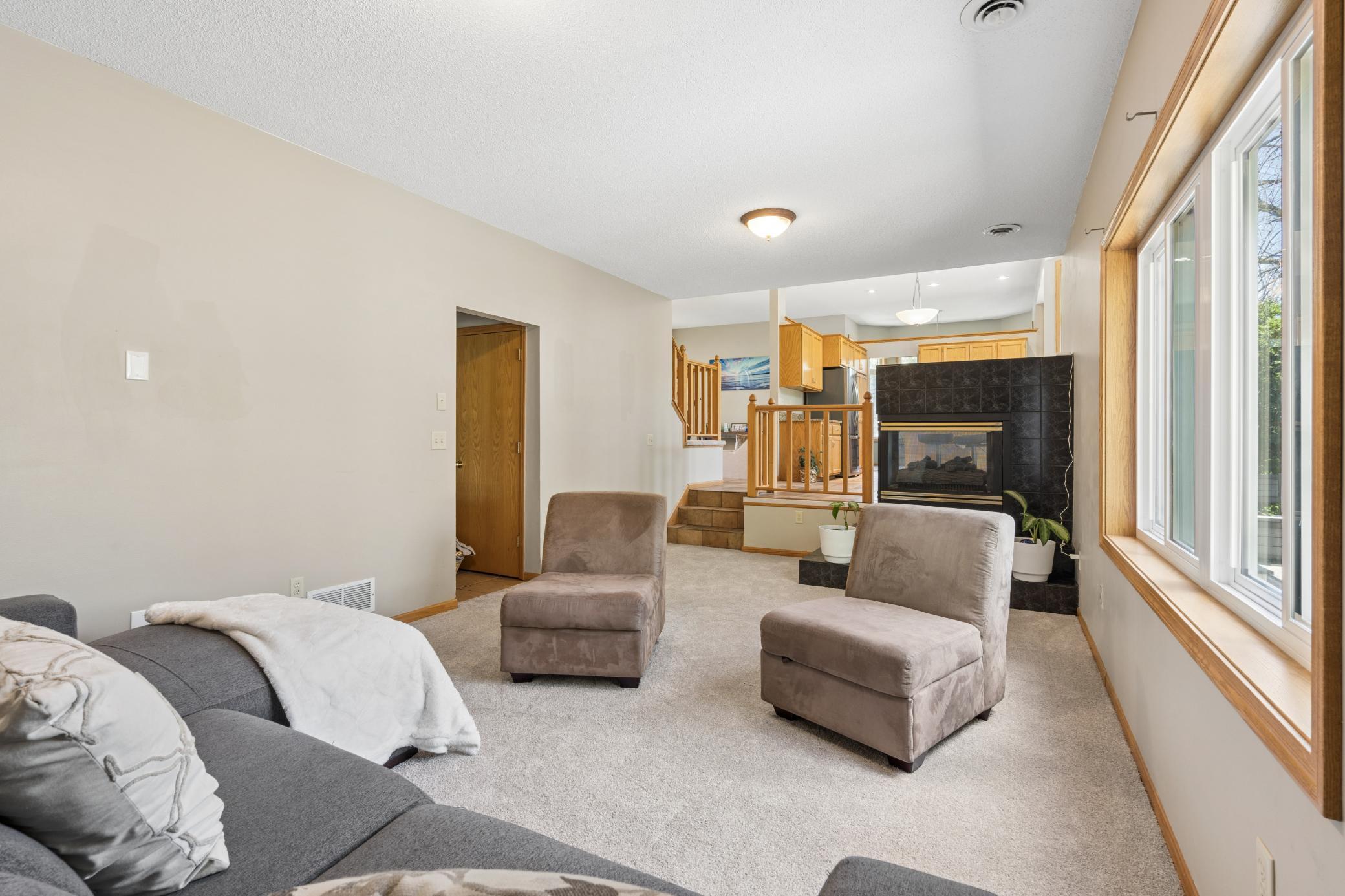 A fireplace anchoring the wall, and a window row that pulls in the yard. The kitchen glows just beyond. Everything you need is already here.